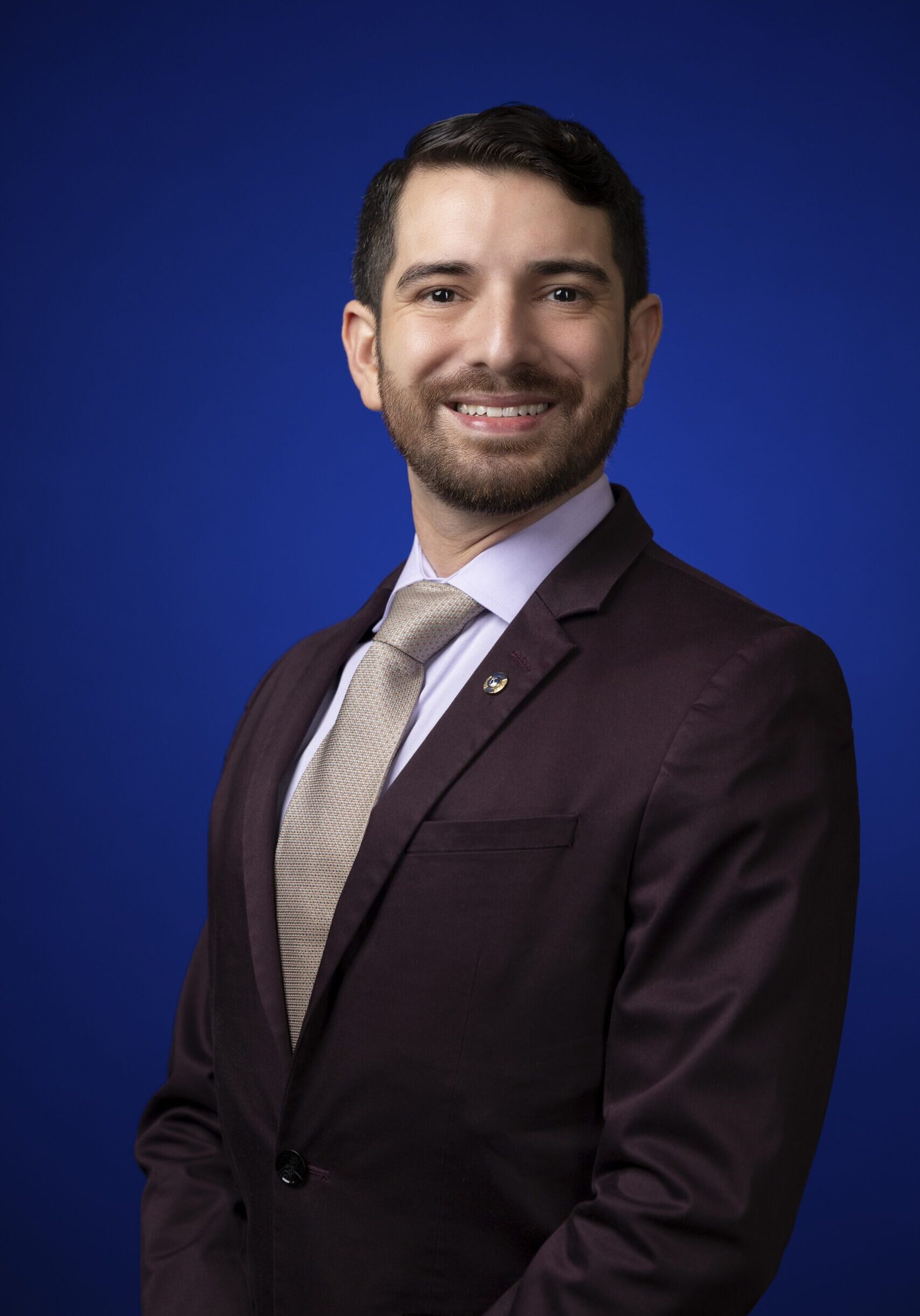 Portrait of 2025-2026 District Governor John Cano. He is in business professional dress with a white shirt, gold tie, and eggplant colored blazer against a blue background.