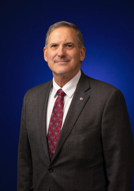 Portrait of 2024-2025 District Governor Bobby Quinten. He is in business professional attire with a white shirt, red tie, and dark gray suit coat against a blue background.